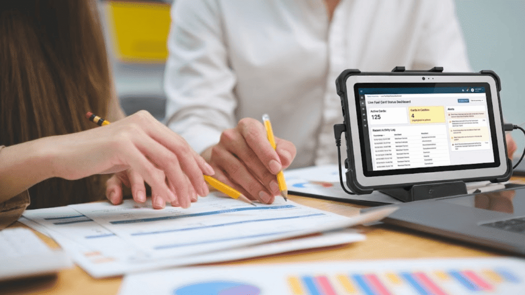 Two people reviewing paperwork with pencils while a tablet displays fuel card data for taxes on a desk next to a fuel card data system.